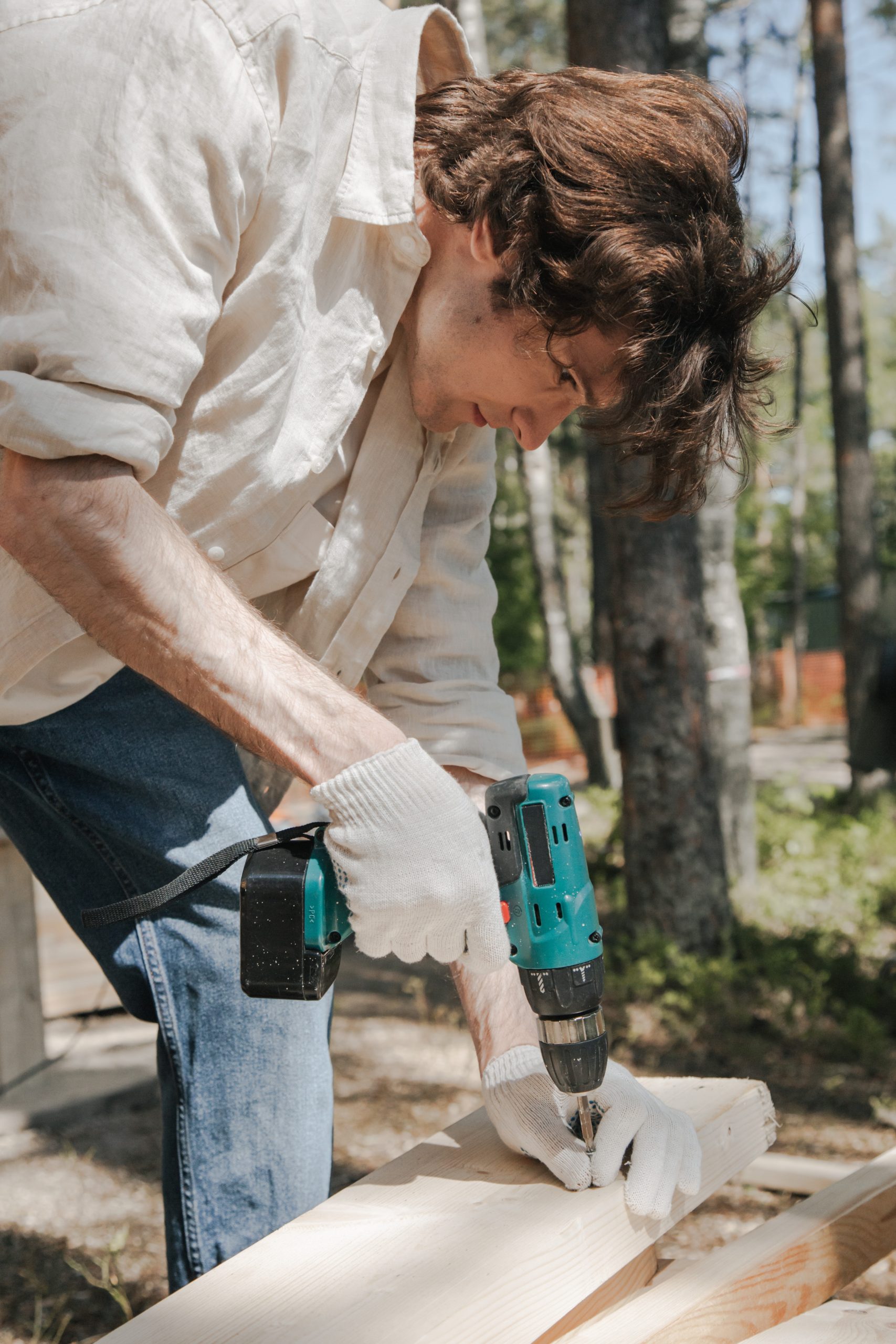 młody mężczyzna wierci dziurę wiertarką akumulatorową w desce, pracując w ogrodzie na świeżym powietrzu
